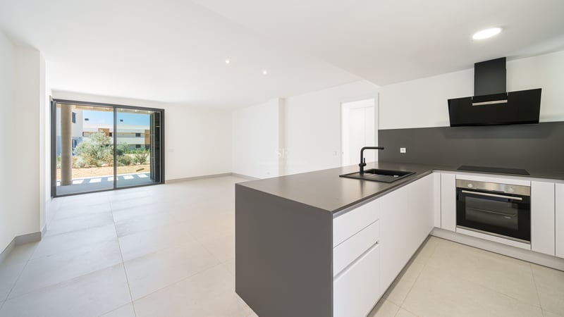 Cocina blanca minimalista con isla gris, electrodomésticos negros y grandes ventanales hacia el exterior.