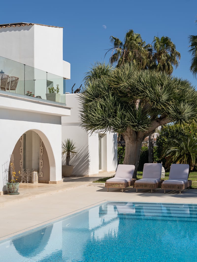 Piscine étincelante à côté d'une villa blanche avec des chaises longues et des palmiers sous un ciel bleu.