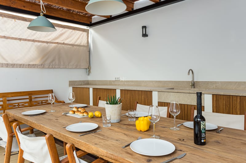 Longue table en bois dressée pour des invités sur une terrasse couverte avec cuisine extérieure.