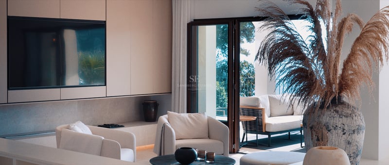 Salon minimaliste lumineux avec fauteuils blancs, grand téléviseur intégré et baies vitrées ouvrant sur une terrasse.