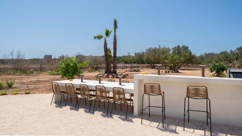 A long white dining table and bar counter with woven chairs on a gravel terrace under a clear blue sky.