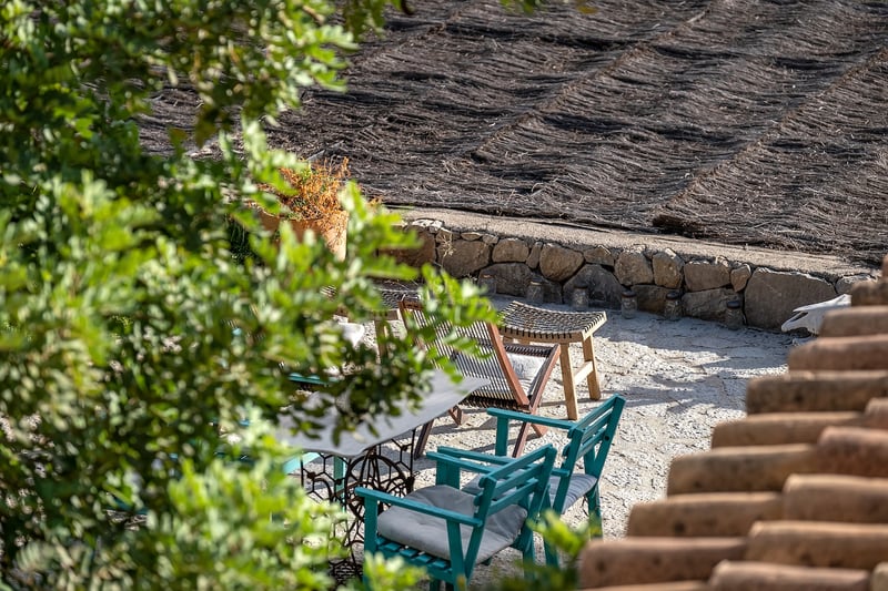 Terrasse en pierre avec chaises en bois bleu sarcelle et toit en chaume, vue à travers la verdure.