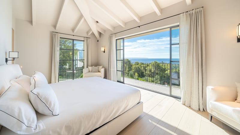 Chambre principale lumineuse avec poutres blanches, parquet en chêne et balcon offrant une vue sur la mer et la forêt.