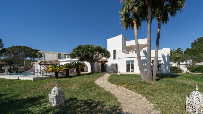 Villa méditerranéenne blanche avec un chemin en pierre, des palmiers et une piscine sous un ciel bleu éclatant.