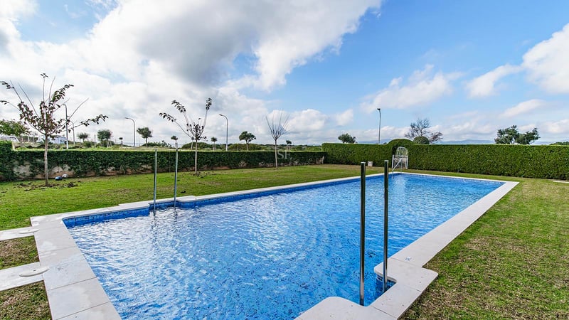 Grande piscine rectangulaire entourée d'une pelouse verte et de haies taillées sous un ciel nuageux.