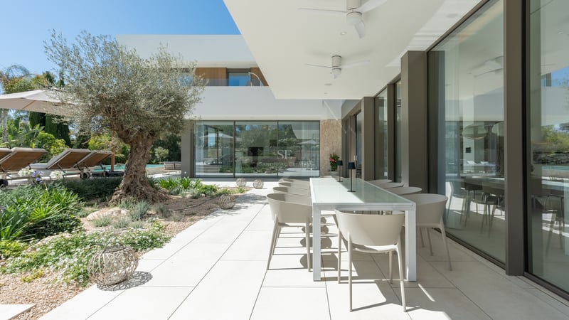 Terrasse de villa contemporaine avec table blanche, grandes baies vitrées et olivier centenaire dans le jardin.