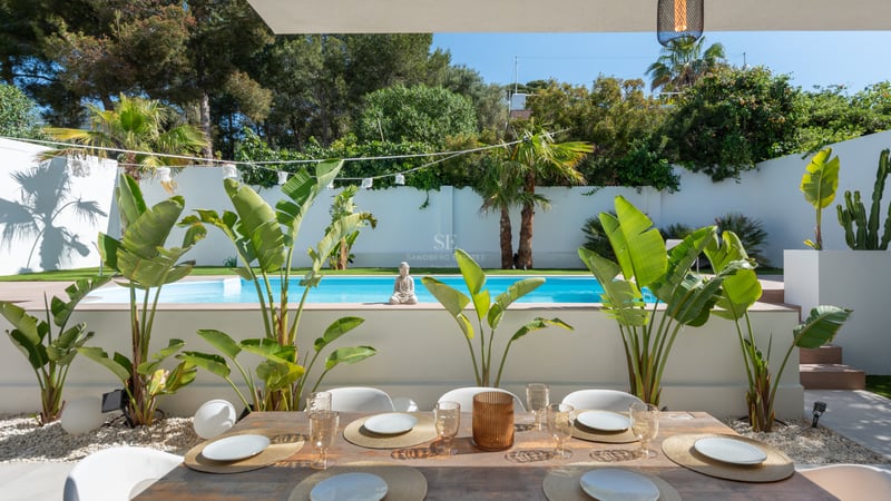 Table de repas en bois devant une piscine turquoise entourée de plantes tropicales et de murs blancs.