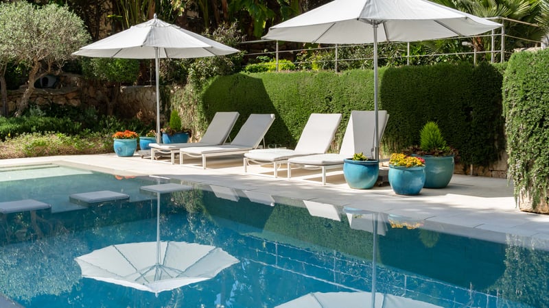 Piscine luxueuse avec quatre chaises longues blanches et parasols assortis devant une haie verte et pots bleus.