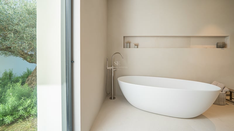 Salle de bain moderne avec baignoire îlot blanche et grande baie vitrée donnant sur un olivier.