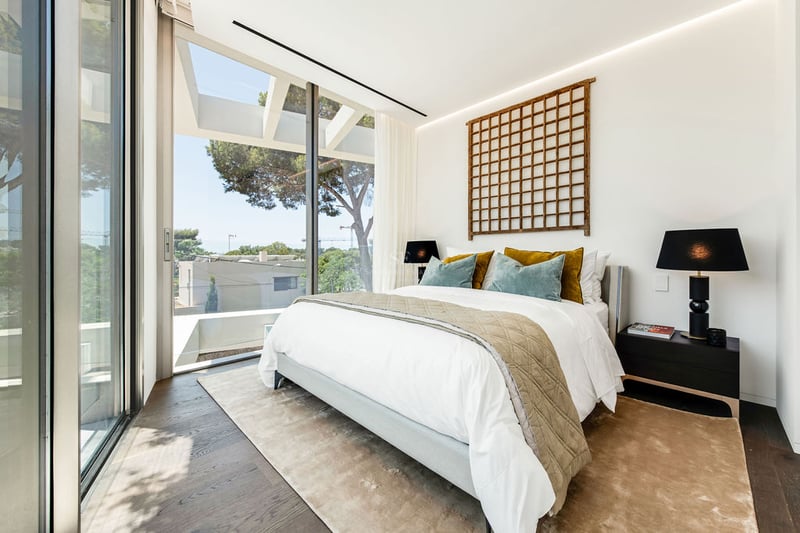 Chambre lumineuse avec parquet foncé, grande baie vitrée et lit blanc orné de coussins en velours bleu et moutarde.