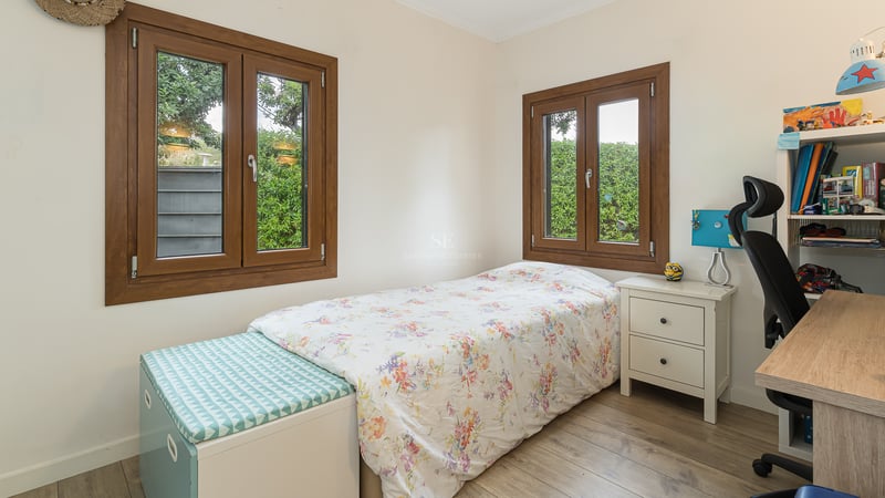 Chambre lumineuse avec un lit simple, un bureau, une chaise de bureau et des fenêtres en bois donnant sur le jardin.
