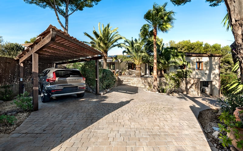 Allée pavée de pierre avec un abri de voiture en bois, des palmiers et une façade de villa en arrière-plan.