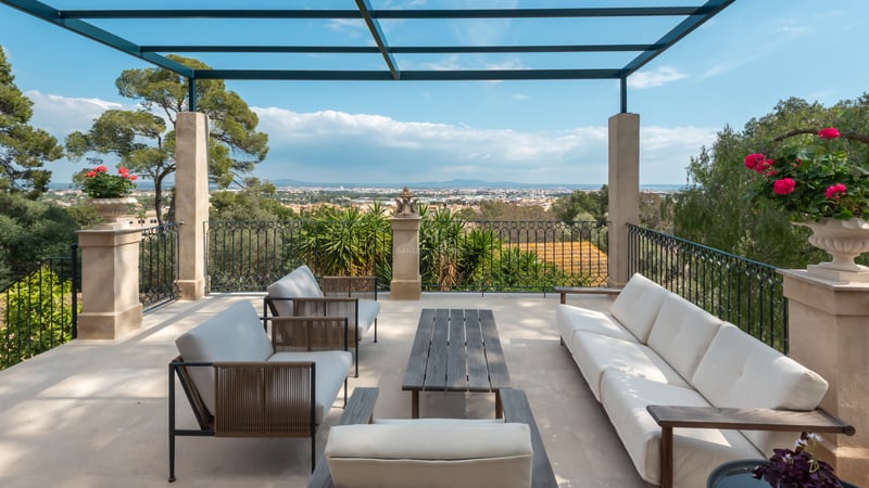 Terrasse de luxe avec canapés blancs, piliers en pierre et balustrade en fer forgé surplombant une vallée et des montagnes.
