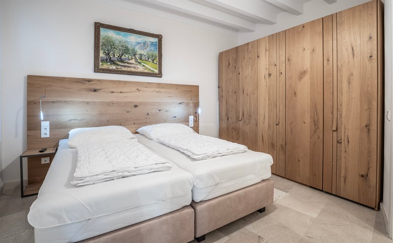 Chambre lumineuse avec plafond à poutres blanches, grand placard en chêne et lits avec tête de lit en bois.