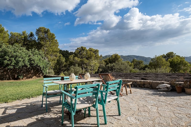 Terrasse en pierre avec table blanche et chaises turquoise entourée de forêts et de montagnes sous un ciel bleu.
