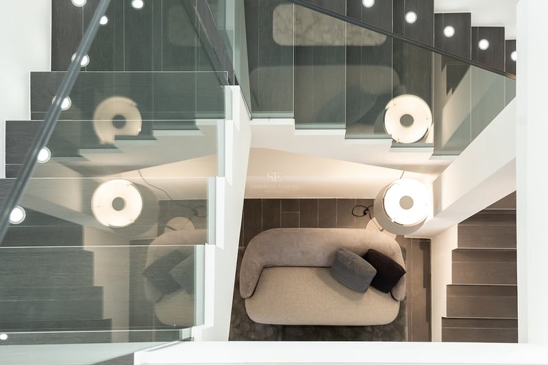 Vue de dessus d'un escalier avec garde-corps en verre donnant sur un canapé gris et une lampe design.
