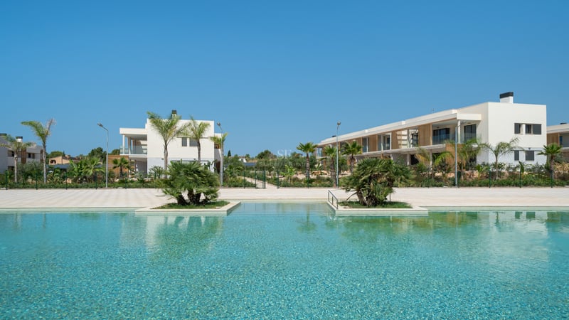 Piscina turquesa con islas de palmeras frente a edificios blancos modernos bajo un cielo azul despejado.