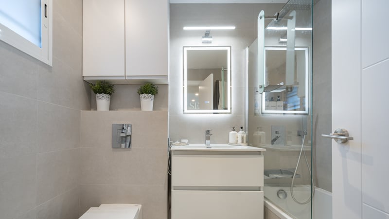 Salle de bain moderne avec vanité blanche, miroir LED, paroi en verre et carrelage beige grand format.