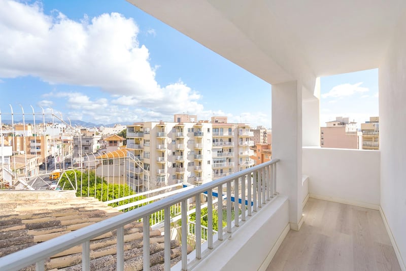 Balcon blanc lumineux avec sol en bois clair surplombant des immeubles urbains et des montagnes au loin.
