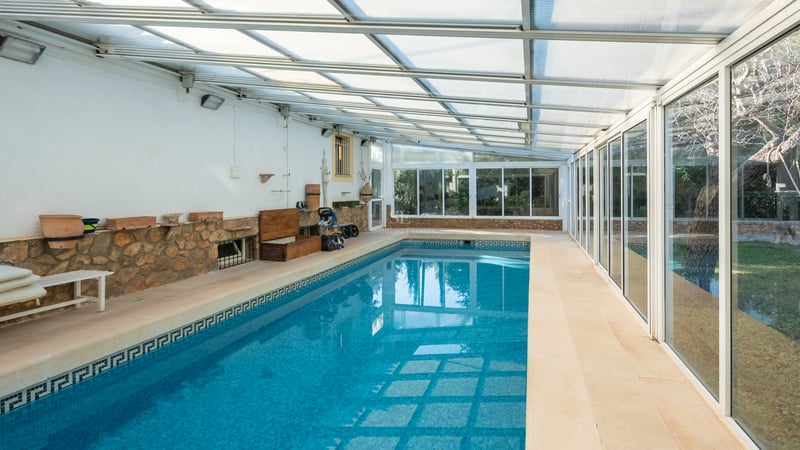 Piscine intérieure avec mosaïques bleues, murs en pierre et toit translucide à côté de grandes fenêtres.