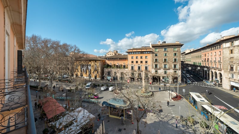 High-angle view from a wrought iron balcony overlooking a bustling historic city plaza with sandstone buildings and trees.