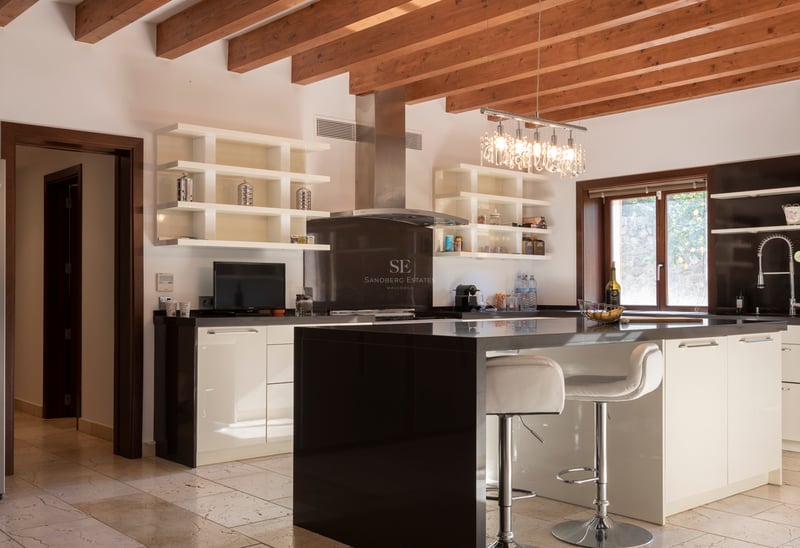 Cuisine moderne avec îlot noir, placards blancs, poutres en bois apparentes et sol en pierre naturelle.