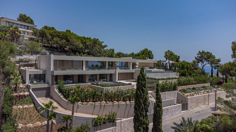 Villa moderne sur plusieurs niveaux sur une colline avec murs en pierre, garde-corps en verre et jardins méditerranéens.