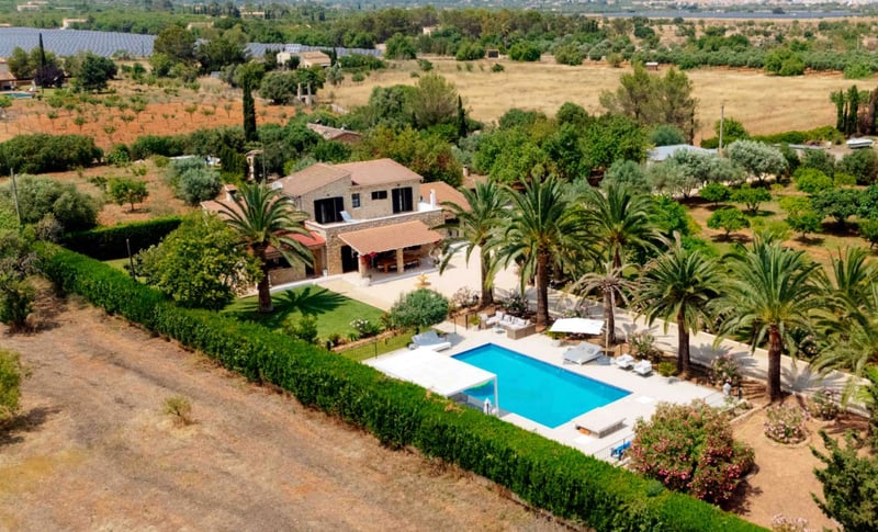 Vue aérienne d'une villa en pierre à deux étages avec piscine bleue, palmiers et grand jardin dans un cadre rural.
