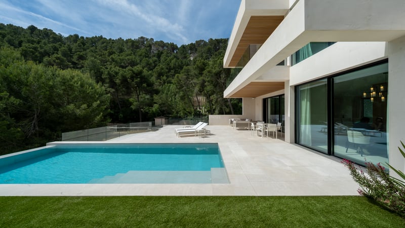 Villa blanche moderne avec piscine turquoise, terrasse en pierre et garde-corps en verre bordant une forêt de pins.