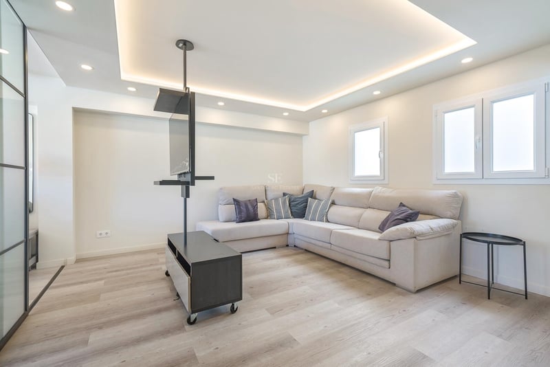 Salon moderne avec grand canapé d'angle beige, parquet clair et support TV pivotant noir minimaliste.