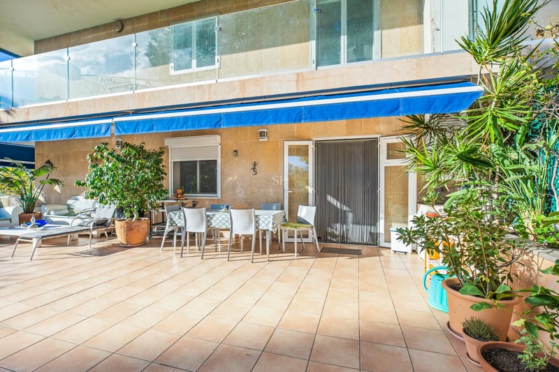 Terrasse ensoleillée avec carrelage, table pour six personnes, stores bleus et balustrades en verre.