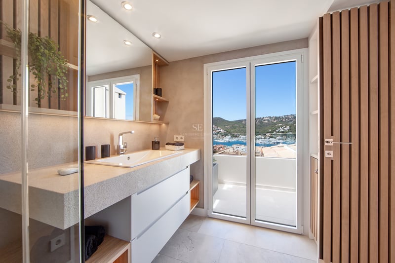 Salle de bain moderne avec vanité en pierre, grand miroir et portes vitrées ouvrant sur un balcon avec vue sur la marina.