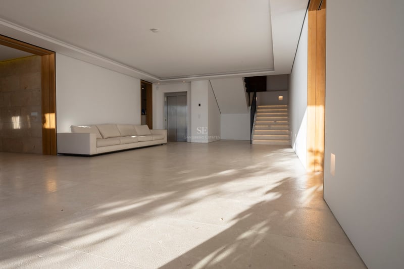 Grand hall minimaliste avec sol en pierre crème, canapé blanc, ascenseur privé et escalier en pierre.