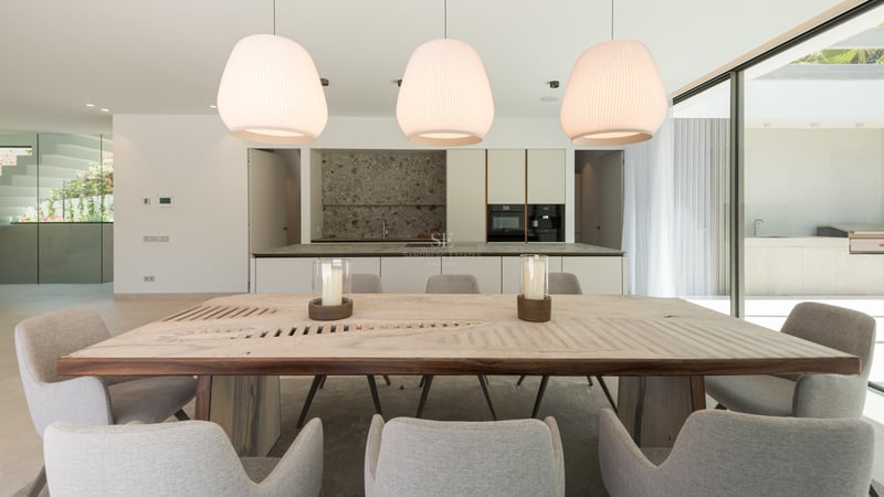 Salle à manger moderne avec table en bois massif, chaises grises et cuisine élégante avec îlot en pierre en arrière-plan.