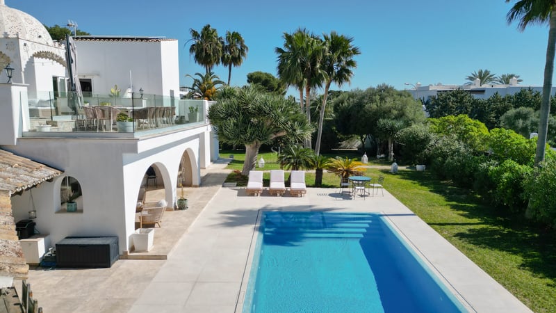 Villa blanche moderne avec piscine rectangulaire bleue, jardin tropical et terrasse avec garde-corps en verre.