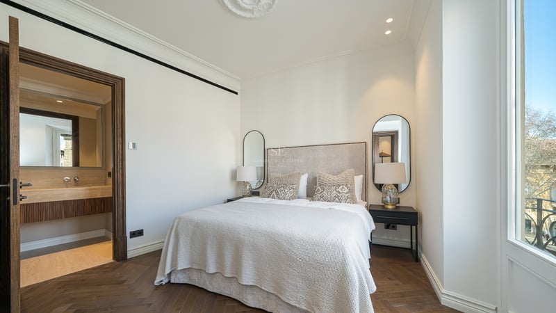 Luxury bedroom featuring oak herringbone floors, a large bed, and a view into a travertine marble en-suite bathroom.