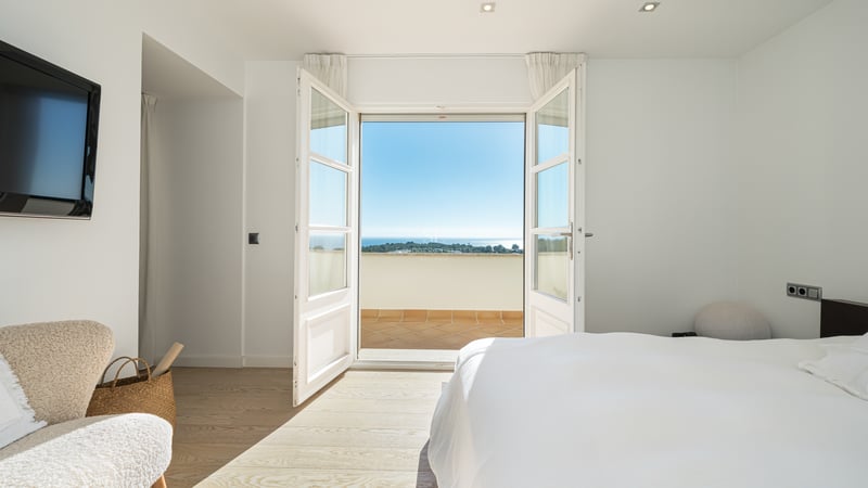 Chambre blanche lumineuse avec portes-fenêtres ouvertes sur une terrasse ensoleillée surplombant la mer.