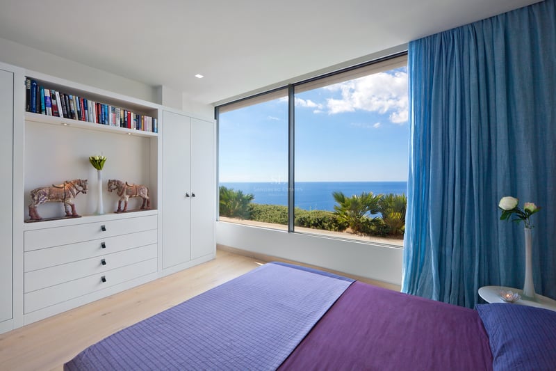 Chambre blanche moderne avec lit violet et grande baie vitrée donnant sur la mer bleue et le ciel.