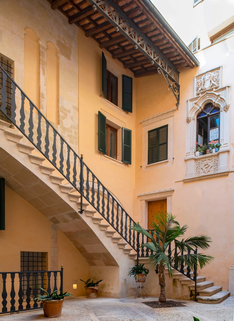 Cour intérieure avec escalier en pierre, rampe en fer forgé, volets verts et un palmier central.