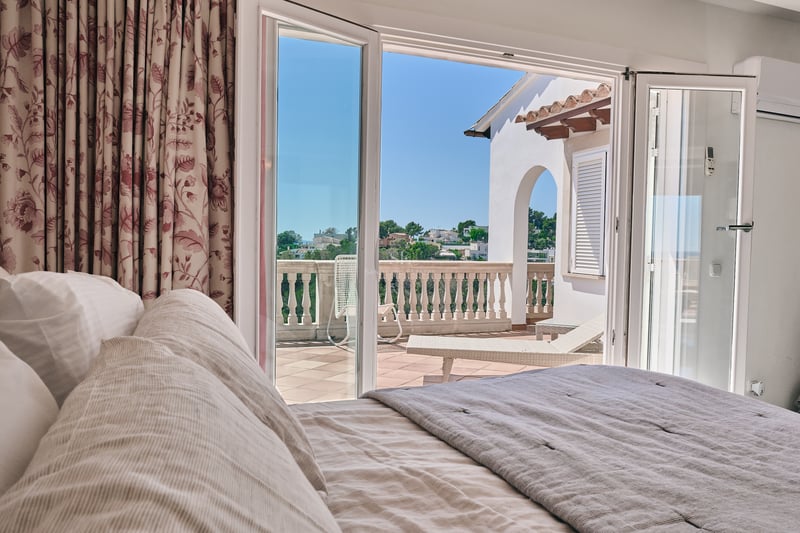 Vue depuis un lit vers une terrasse ensoleillée par des portes vitrées ouvertes avec une balustrade en pierre.