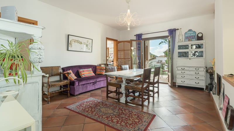 Salle à manger avec sol en terre cuite, canapé en velours violet et table en bois blanc ouvrant sur un balcon.