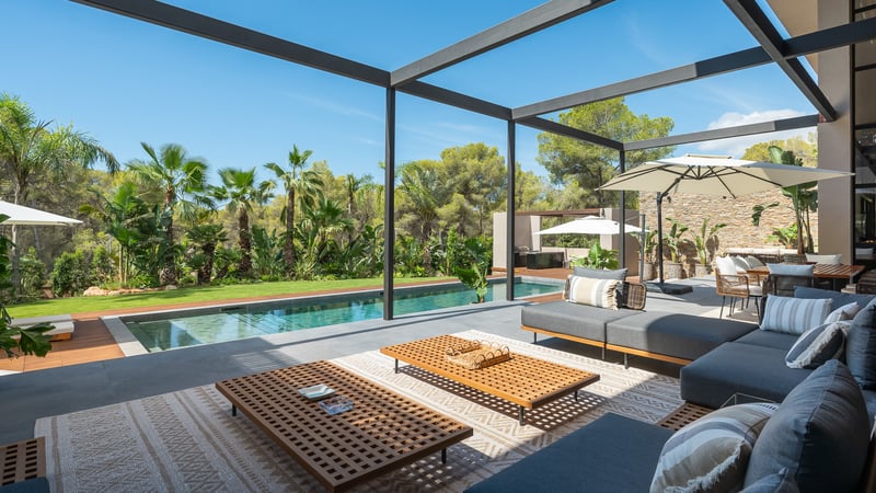 Terrasse moderne avec canapés gris, tables en bois et piscine face à un jardin tropical luxuriant.