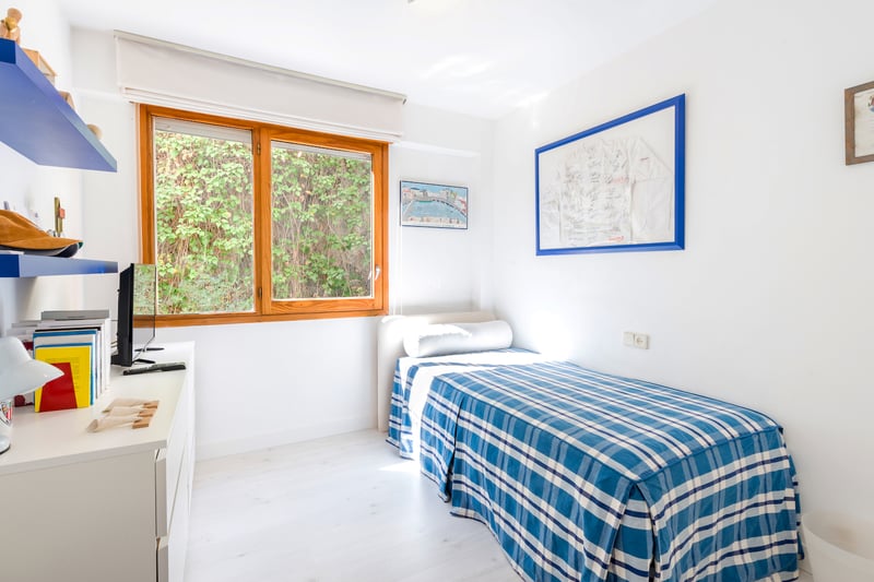 Chambre simple lumineuse avec un lit à carreaux bleus, un bureau blanc et une grande fenêtre en bois.