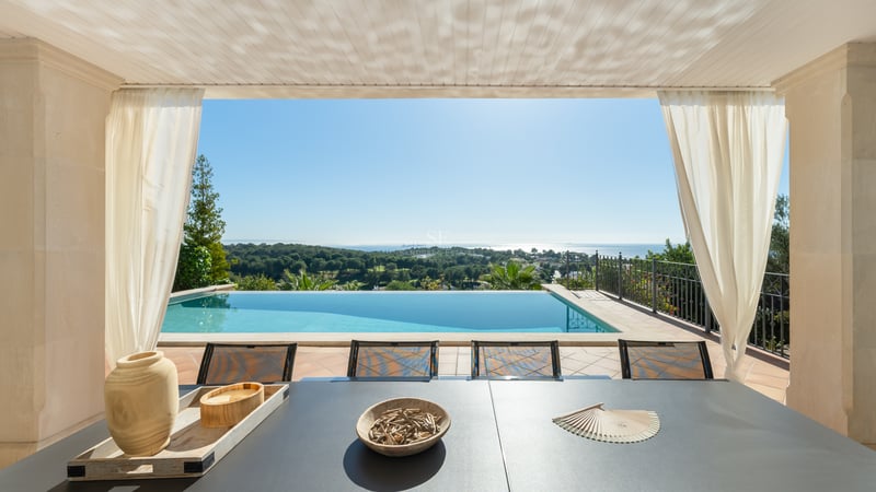 Terrasse ombragée avec table à manger surplombant une piscine à débordement, une pinède et la mer Méditerranée.
