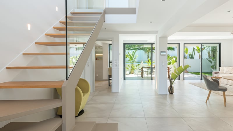 Couloir moderne avec escalier suspendu en bois et garde-corps en verre, s'ouvrant sur le salon et le jardin.