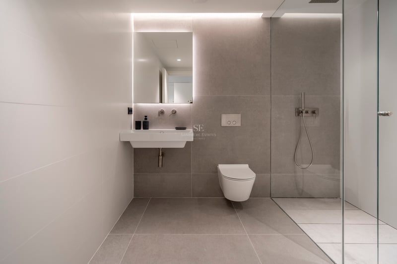 Salle de bain moderne avec carrelage gris, miroir rétroéclairé, lavabo blanc, WC suspendu et paroi de douche en verre.