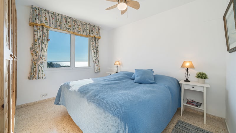 Une chambre lumineuse avec un dessus de lit bleu, vue sur la mer par une fenêtre aux rideaux fleuris et lampes de chevet.