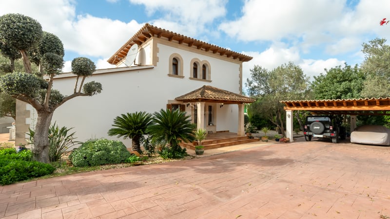 Villa blanche à deux étages avec toit en terre cuite, fenêtres cintrées et une large allée avec parking couvert.