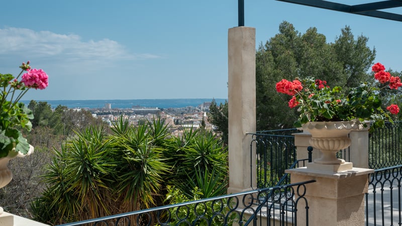 Vue depuis une terrasse sur une ville méditerranéenne et la mer, avec fleurs rouges et balustrade en fer forgé.