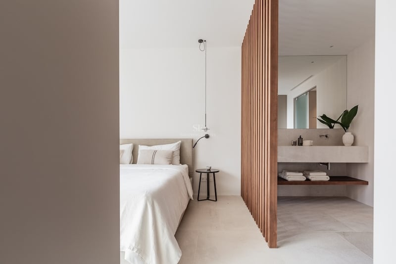 Chambre moderne avec lit beige et cloison en bois séparant un espace salle de bain en pierre.